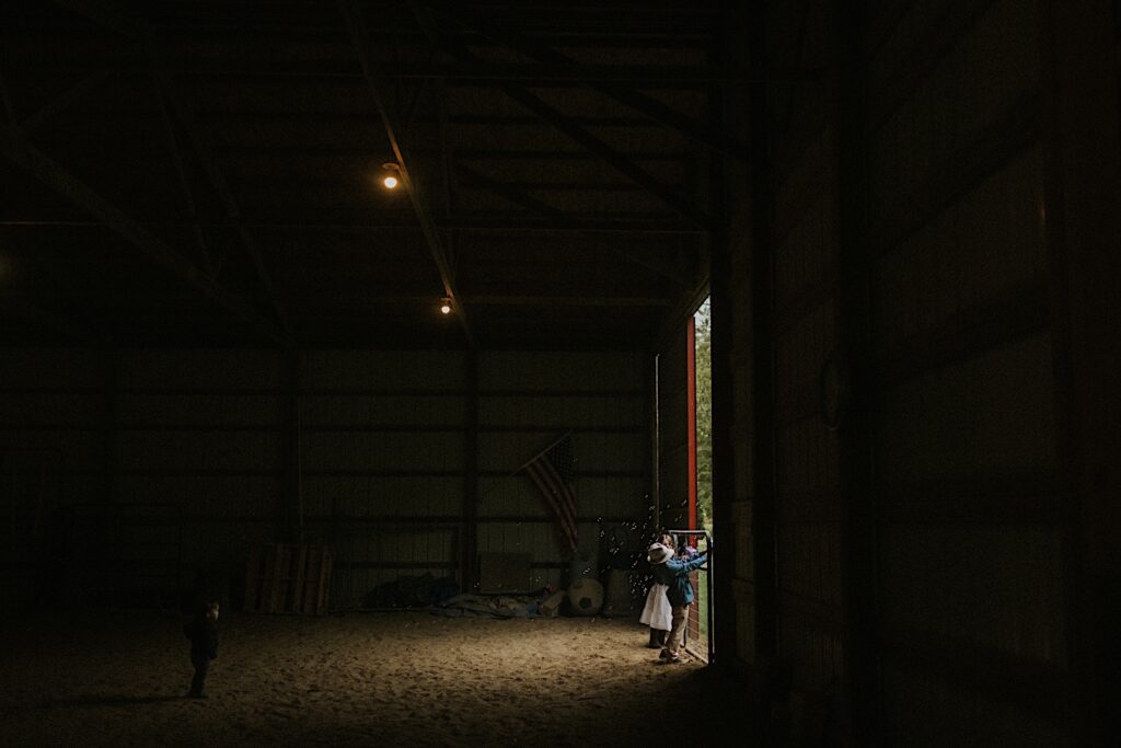 Two kids stand at the entrance of a barn and shoot bubbles out of a bubble gun through the open door