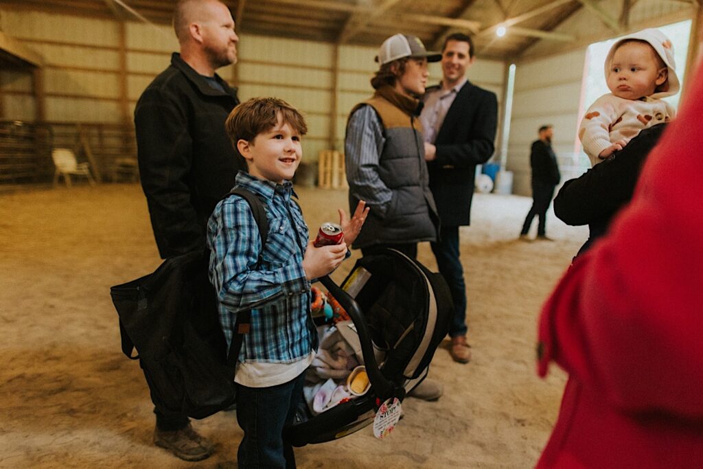 A young boy holds a can of coke and stands around other guests of a wedding ceremony that took place inside a barn