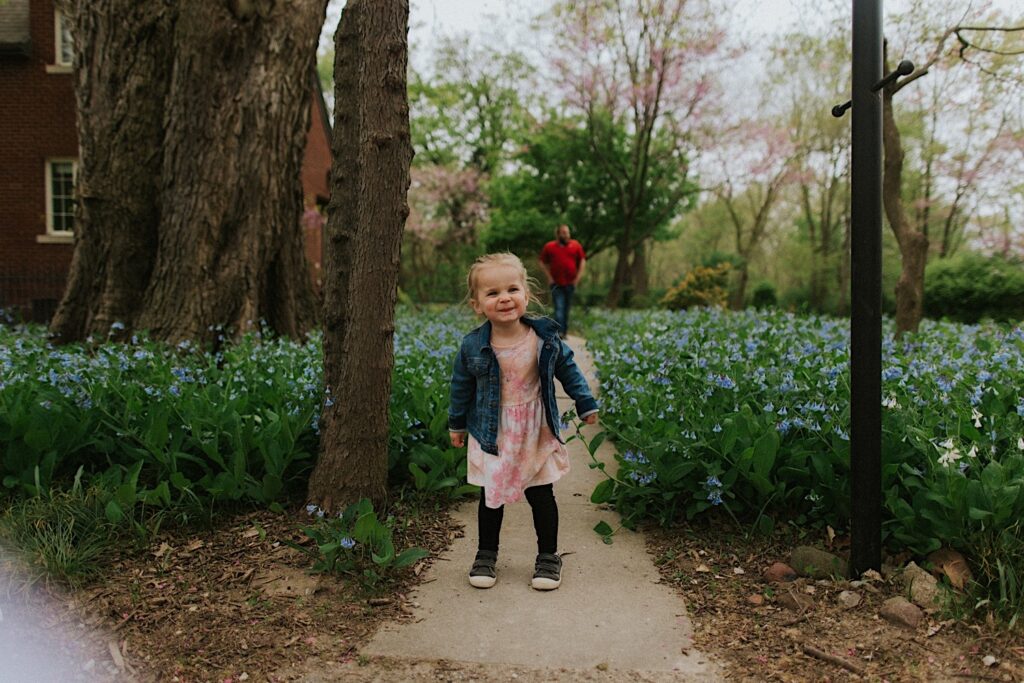 A young girl makes a funny face to the camera as she stands on a sidewalk in a backyard with blue flowers on either side of it