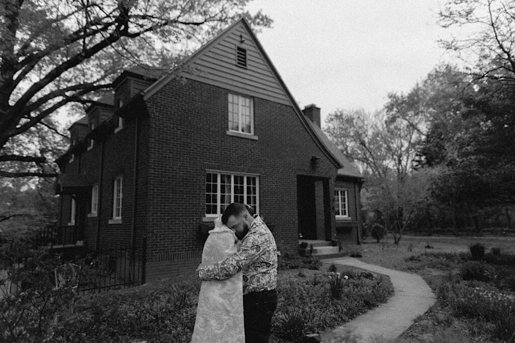 Black and white photo of a bride in a white blanket being hugged by the groom outside their Springfield home after their backyard barn wedding