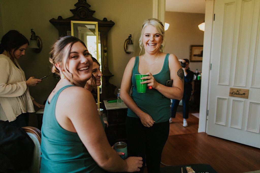 Three bridesmaids gather in a getting ready space and smile at the camera with drinks in hand before a wedding