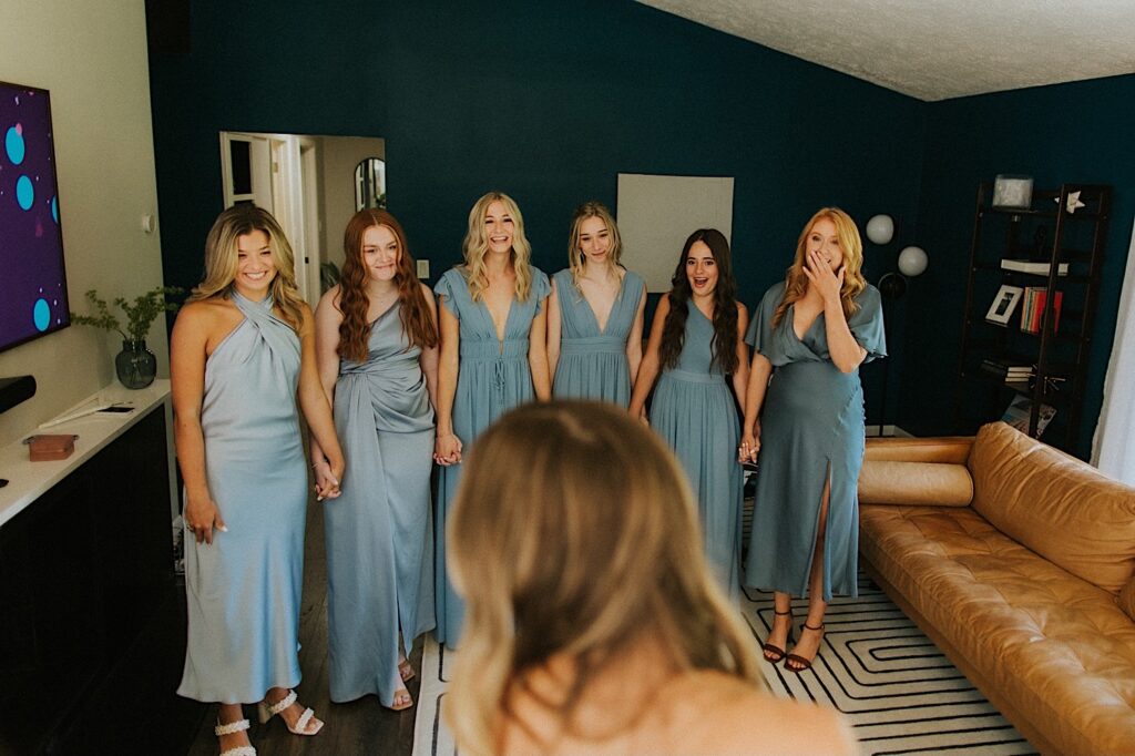 Six bridesmaids hold hands and smile during a first look with the bride in her wedding dress who is in the foreground of the photo