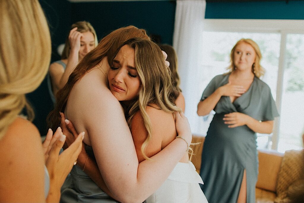 A bride hugs a bridesmaid and starts to tear up on her wedding day as the other bridesmaids all stand around her