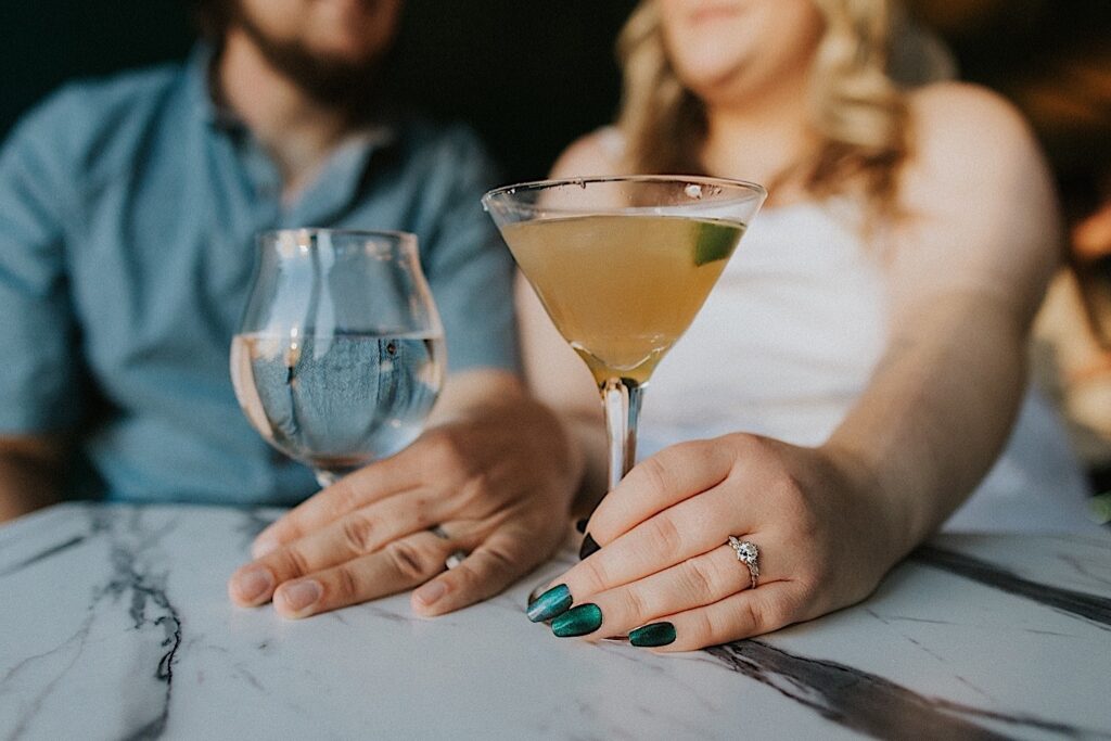 Detail photo of 2 drinks on a table being held by a couple in the background, the woman is wearing an engagement ring
