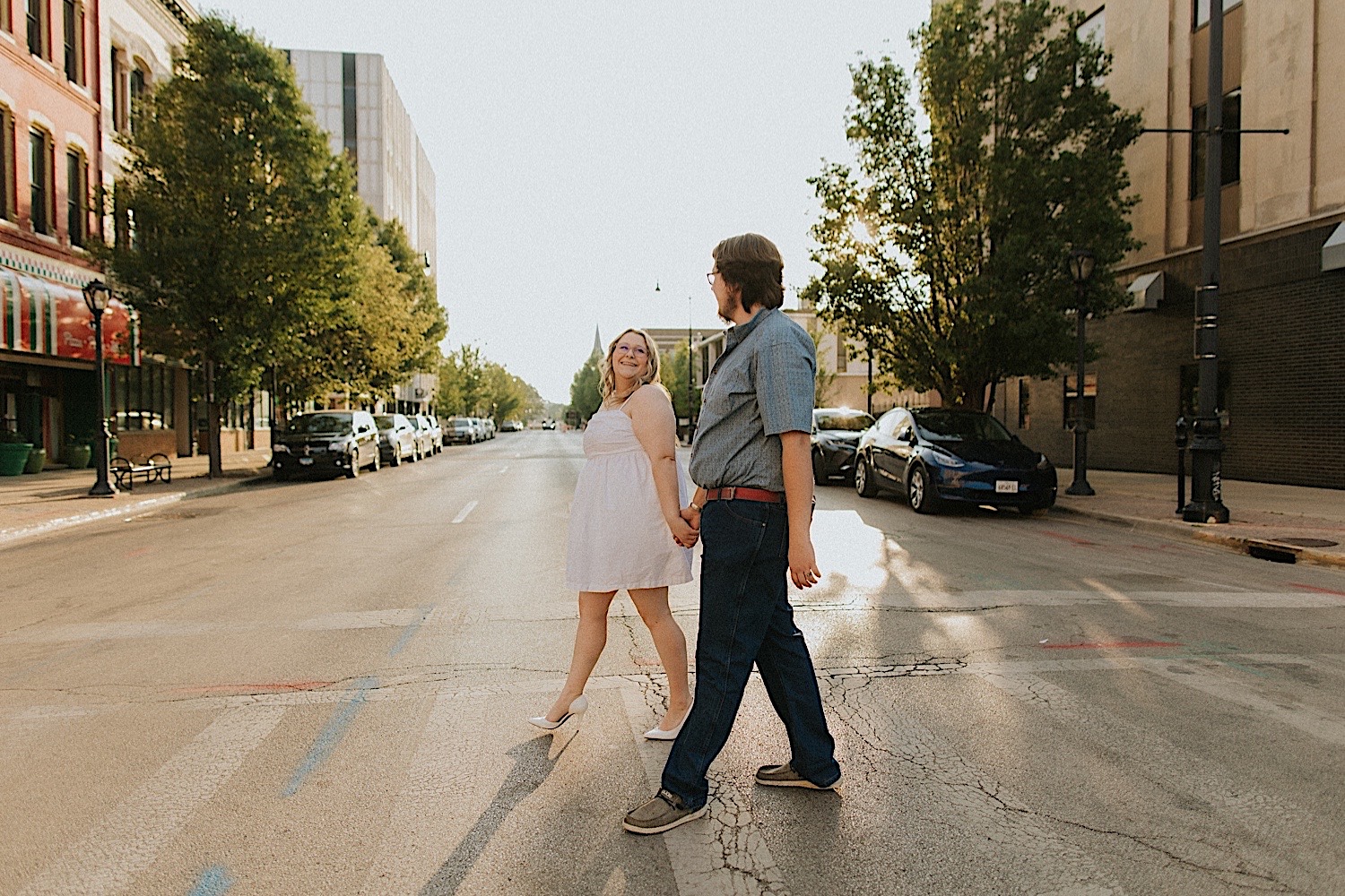 A couple walk hand in hand and smile at one another while crossing a street in downtown Springfield during their engagement session