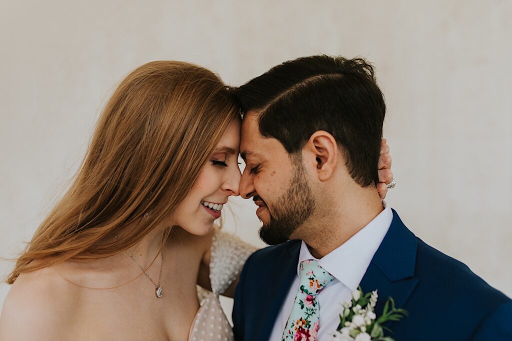 A bride and groom smile at one another with their eyes closed while touching their foreheads together