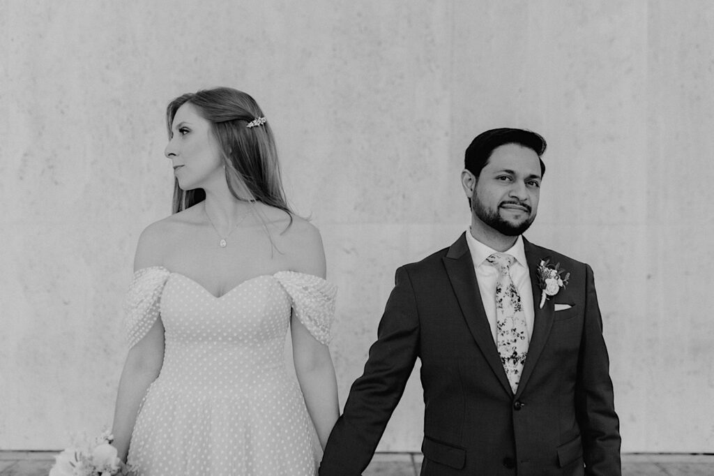 Black and white photo of a bride and groom standing side by side and holding hands, the bride is looking to the left while the groom looks at the camera