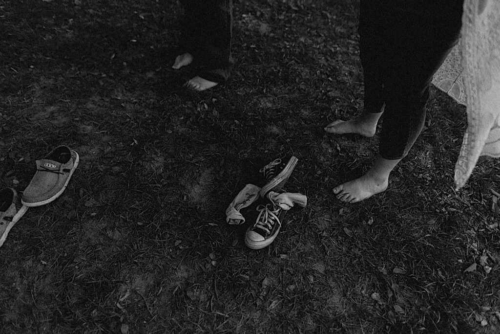 Black and white photo of 2 pairs of shoes on the ground with a pair of bare feet next to them