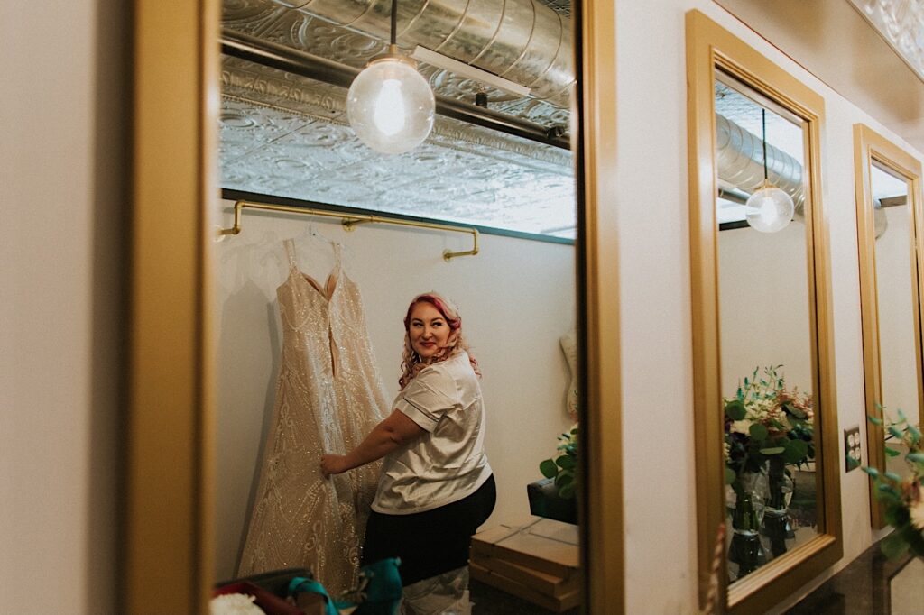 A woman holds her wedding dress that is hanging on the wall in front of her and looks over her shoulder at a mirror on the wall