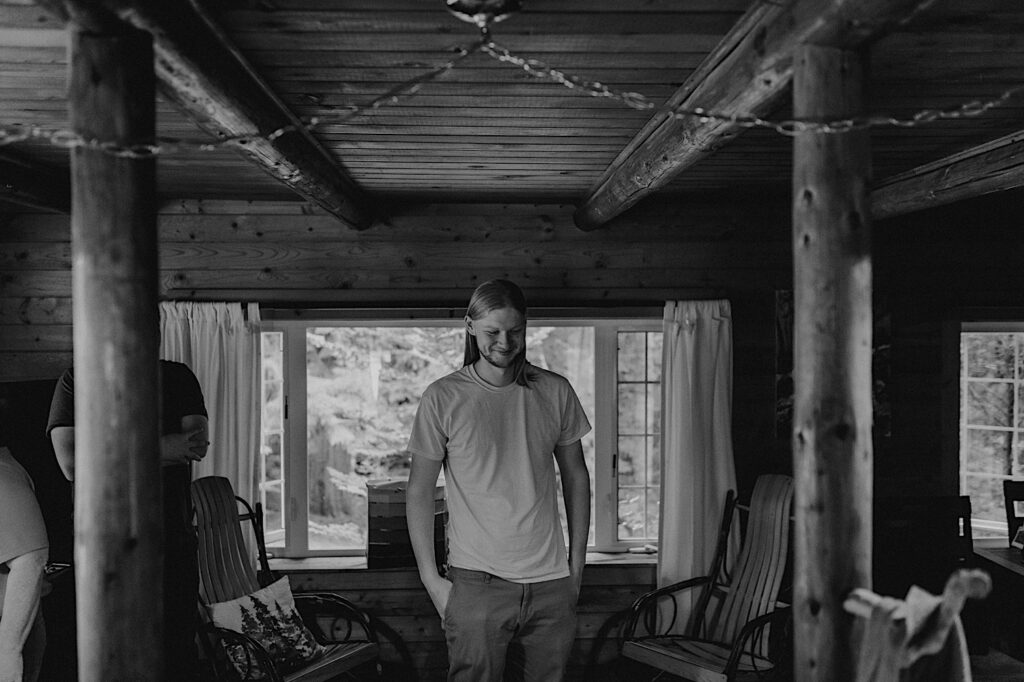Black and white photo of a man smiling in the middle of a room in what looks like a log cabin