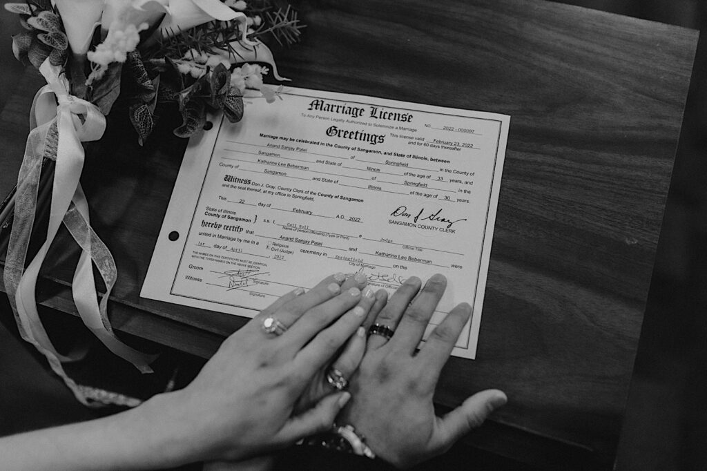 Black and white photo of a marriage license signed with the bride and grooms hands touching it