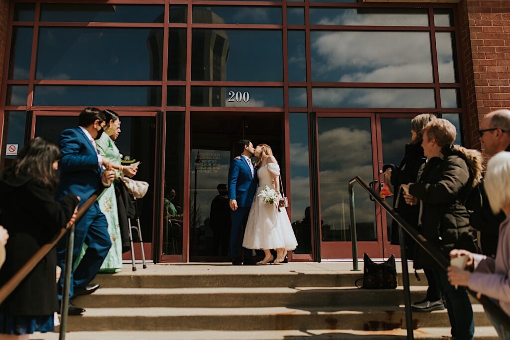 A bride and groom kiss one another while exiting the Sangamon County Courthouse after their elopement ceremony as their families line either side of the staircase in front of them
