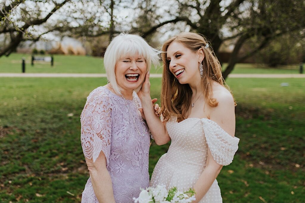 A bride smiles and touches her mother's face who is also smiling as the two stand in a park together after the bride's elopement at the Sangamon County Courthouse