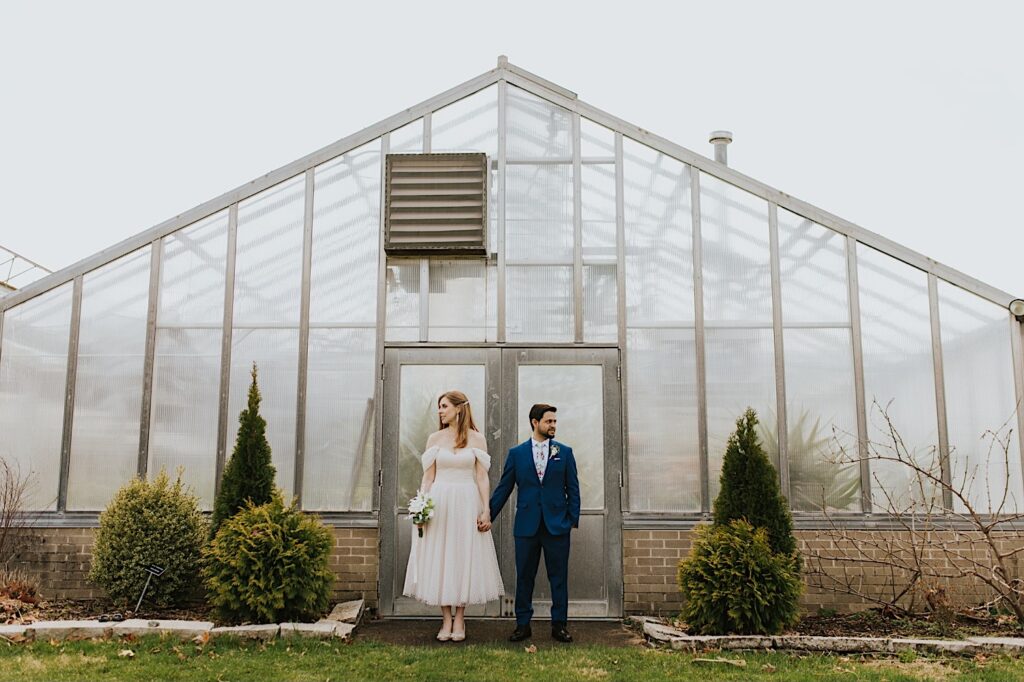 A bride and groom stand next to one another and hold hands while looking in opposite directions standing outside of a greenhouse after their elopement at the Sangamon County Courthouse