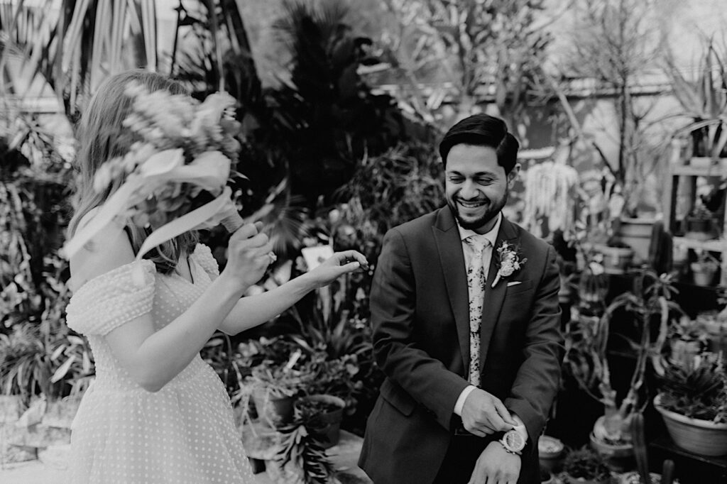 Black and white photo of a bride messing with a groom as he smiles while they stand in a greenhouse after their elopement at the Sangamon County Courthouse