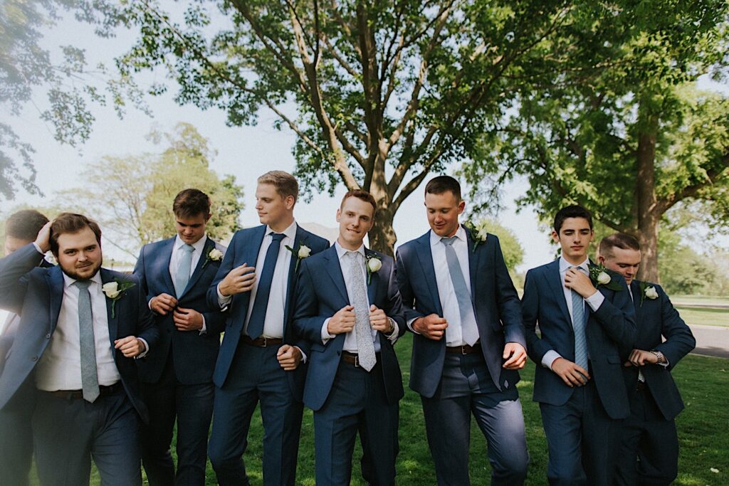 A groom stands with his 7 groomsmen in a park before his wedding at Bloomington Country Club, the groom is smiling at the camera while the other groomsmen adjust their suit coats
