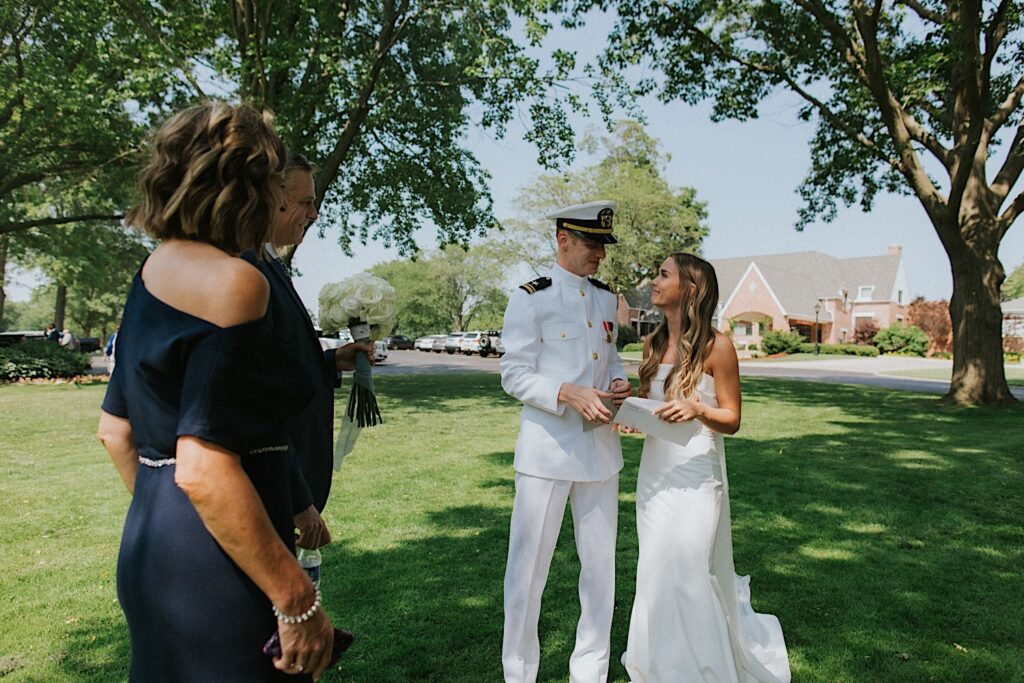 A bride and groom in his military uniform stand next to one another and smile with the bride's parents in front of them as they stand in a park outside the Bloomington Country Club before their wedding ceremony