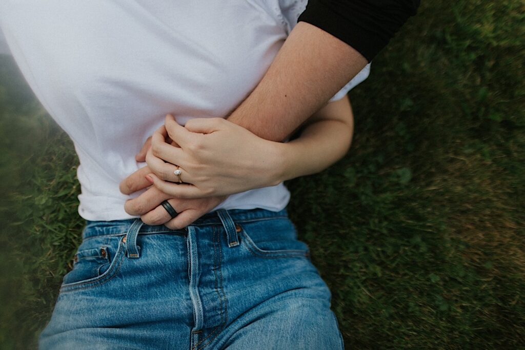 Close up photo of a woman laying on the grass, her hand with an engagement ring on it is holding the hand of a man which also has an engagement ring on his hand