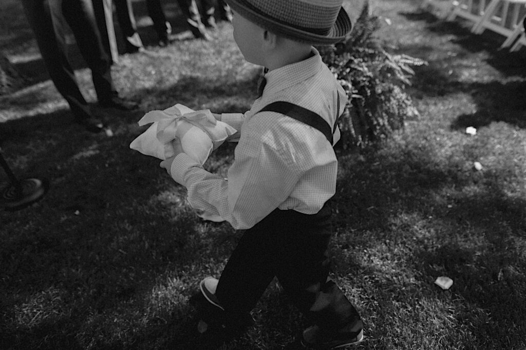 Black and white photo of a young boy carrying the rings of a wedding ceremony down the aisle of a wedding ceremony
