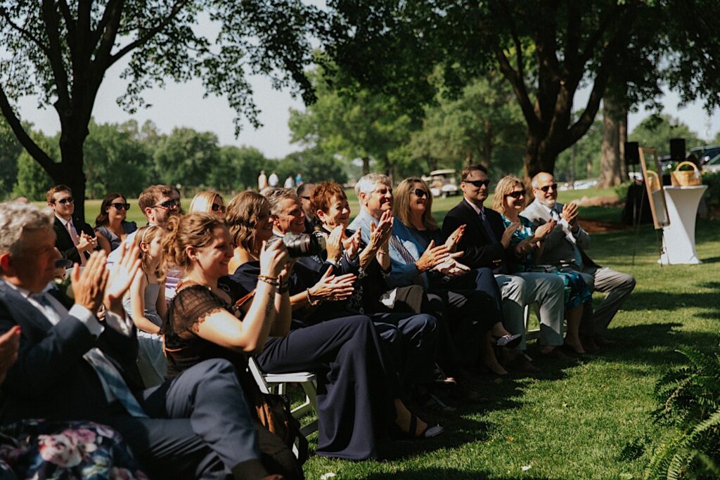 Guests of an outdoor wedding ceremony at Bloomington Country Club clap and cheer towards the bride and groom who are off camera