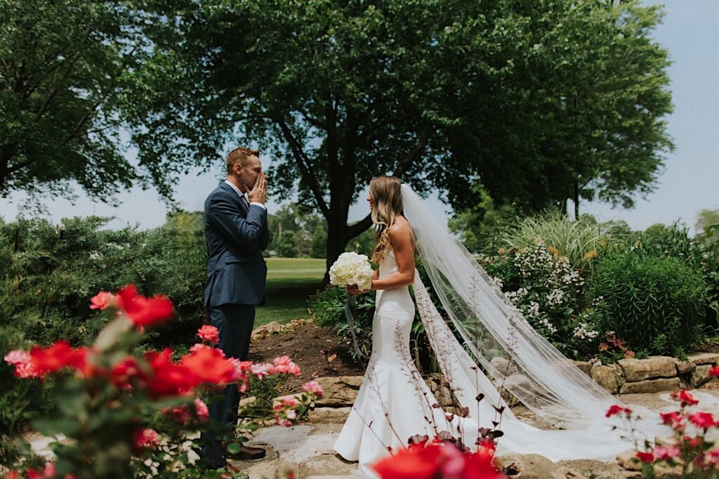 A groom covers his mouth as he sees his bride for the first time while the two stand together in a park
