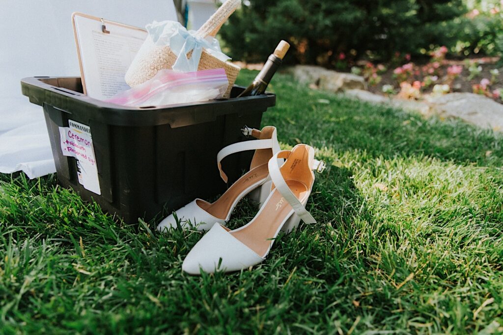 A brides shoes sit in the grass next to a tote filled with wine bottles and other items