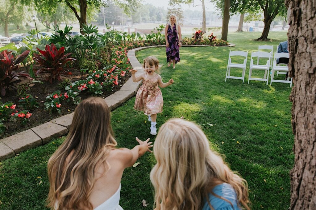 A young child runs towards a bride in a park as the bride crouches down and extends her arms out towards the child to catch her