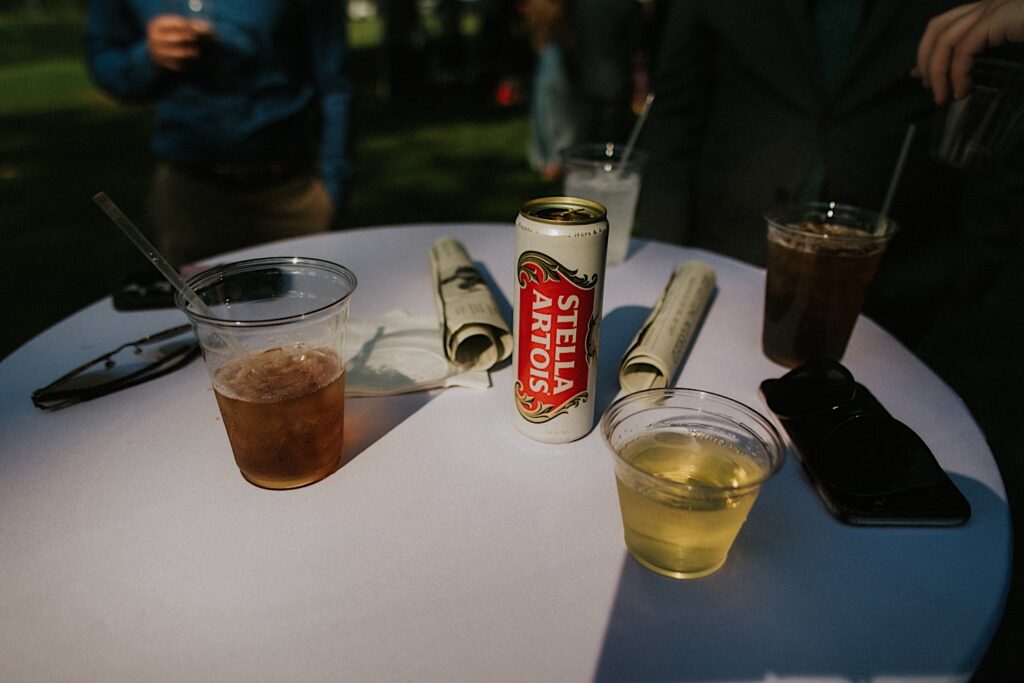 A can of Stella Artois sits on a table with other drinks with guests of a wedding standing around the table