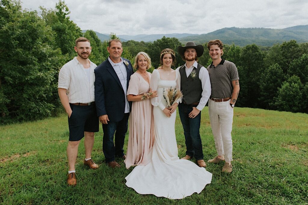 A bride and groom stand with family members on a hillside overlooking a forest and hills while all smiling at the camera