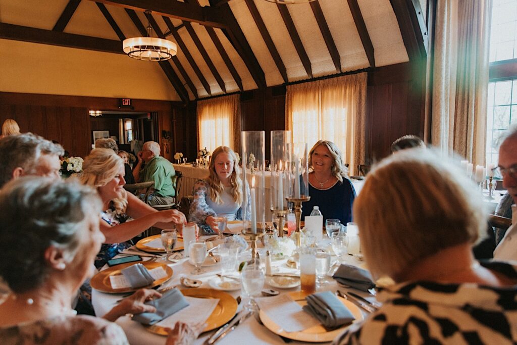Guests of an indoor wedding reception at the Bloomington Country Club sit at a table and mingle with one another before dinner