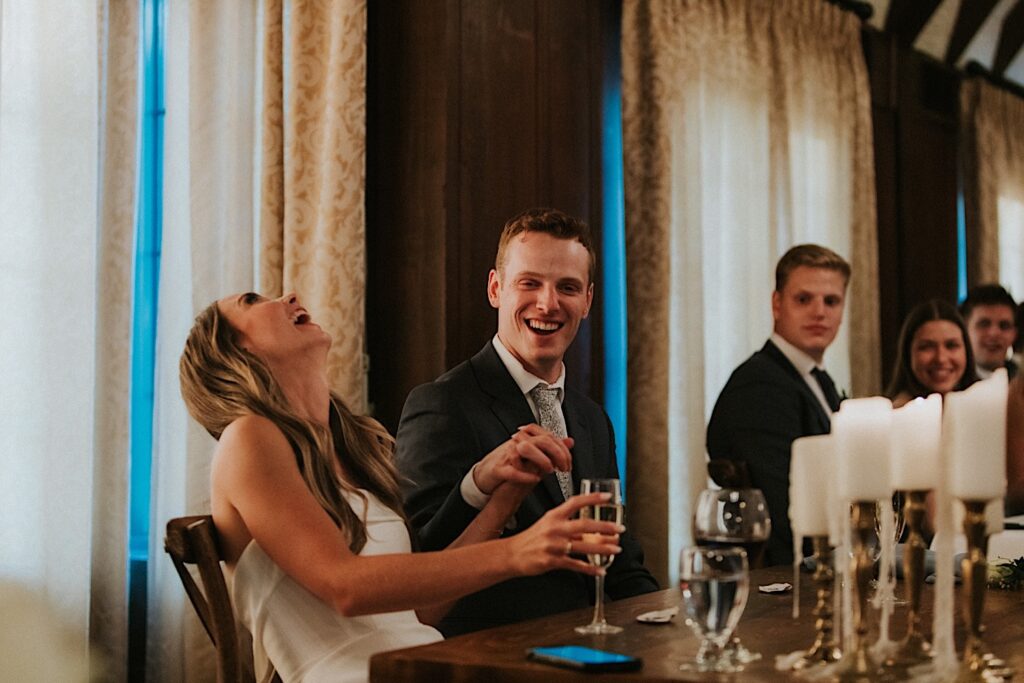 A bride leans back and laughs as she holds the grooms hand who is sitting next to her during their wedding reception at Bloomington Country Club
