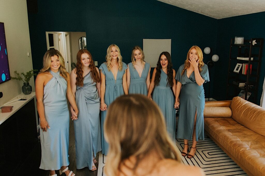 A group of 6 bridesmaids stand side by side holding hands and smiling as the bride in the foreground stands facing them during her first look