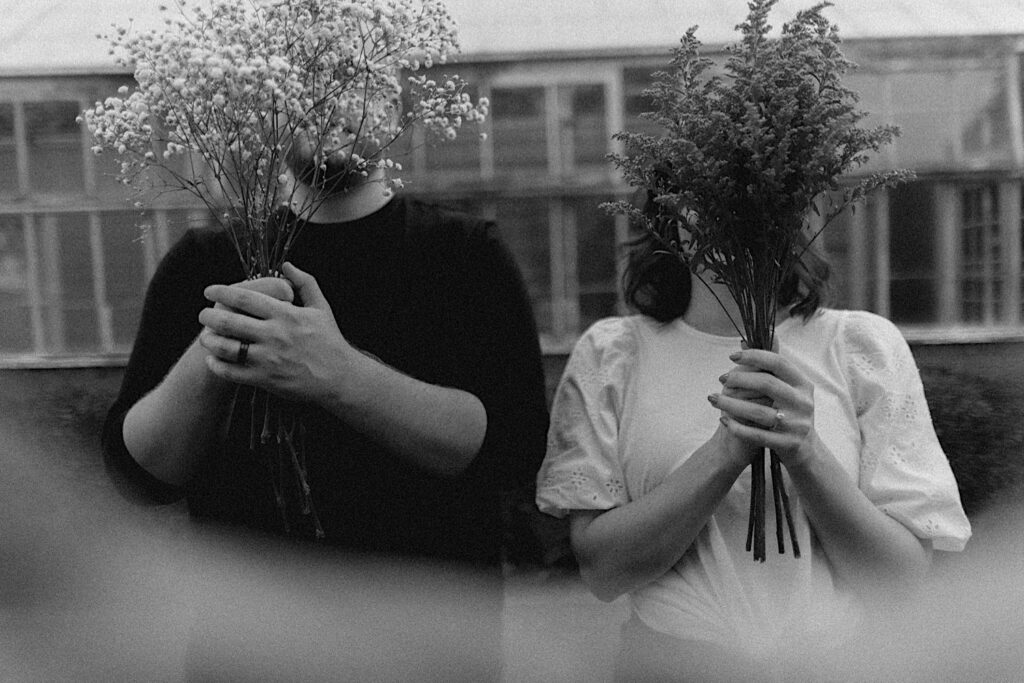 During their engagement session at Allerton Park, a couple stand in front of a greenhouse with bouquets of flowers held in front of their faces, the photo is in black and white