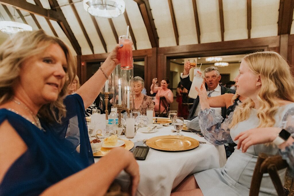 Guests of a wedding reception at Bloomington Country Club sitting at a table lift their glasses during a speech