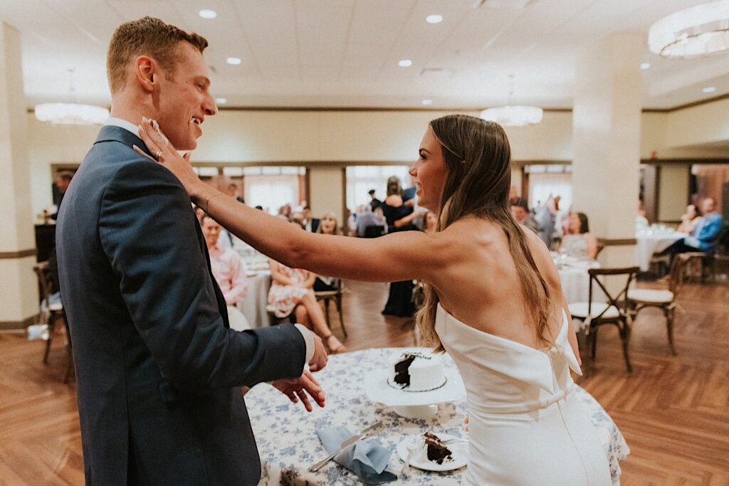 At a wedding reception at the Bloomington Country Club a bride smears cake on a grooms face as he smiles and guests watch