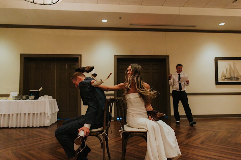 At their wedding reception at the Bloomington Country Club a bride and groom sit back to back to play the shoe game, the bride is turning around to touch the groom's shoulder as he laughs