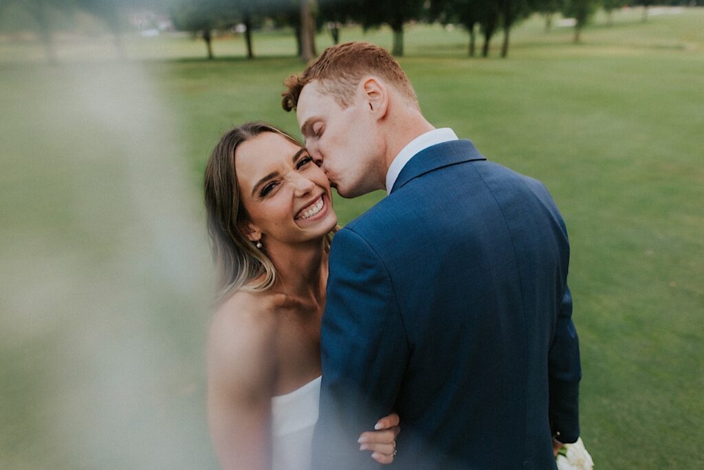 A bride smiles at the camera as the groom kisses her on the cheek during their wedding at the Bloomington Country Club, they're standing on the golf course in the photo