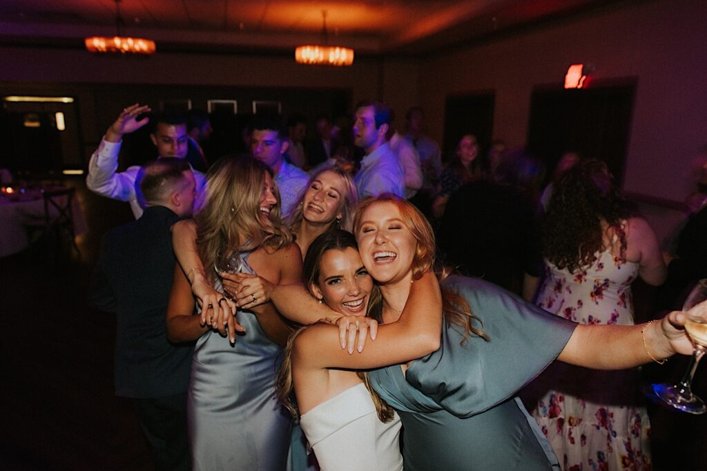 A bride smiles at the camera while hugging one of her bridesmaids on the dancefloor with other guests behind them during their indoor wedding reception at the Bloomington Country Club