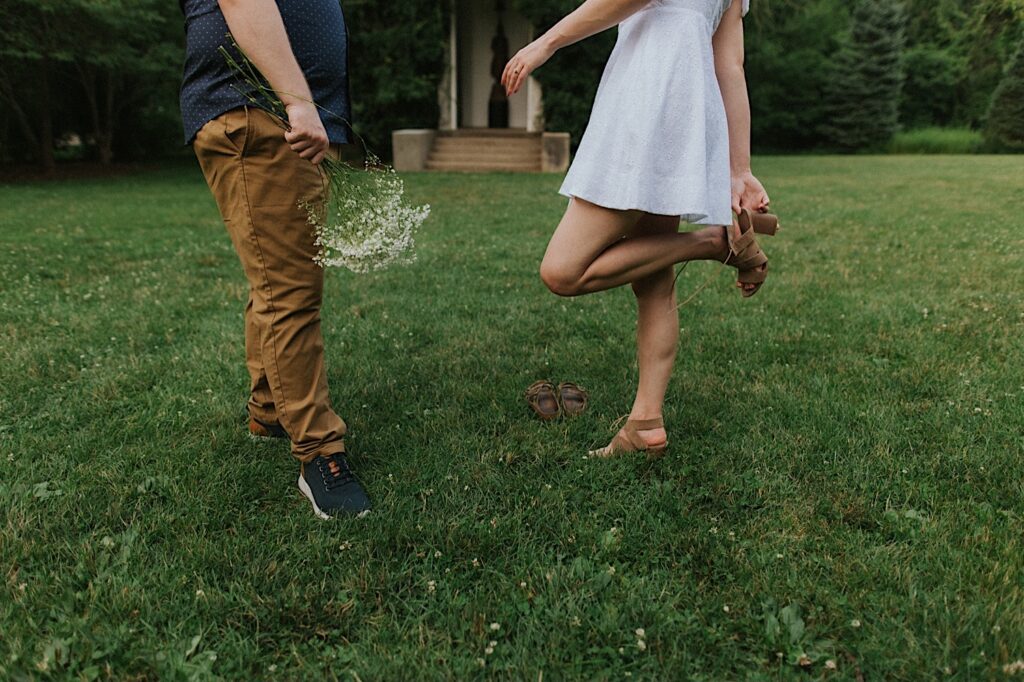 Waist down photo of a man and woman standing in a field during their engagement session at Allerton Park, the man is holding a flower bouquet while the woman is swapping the shoes that she is wearing