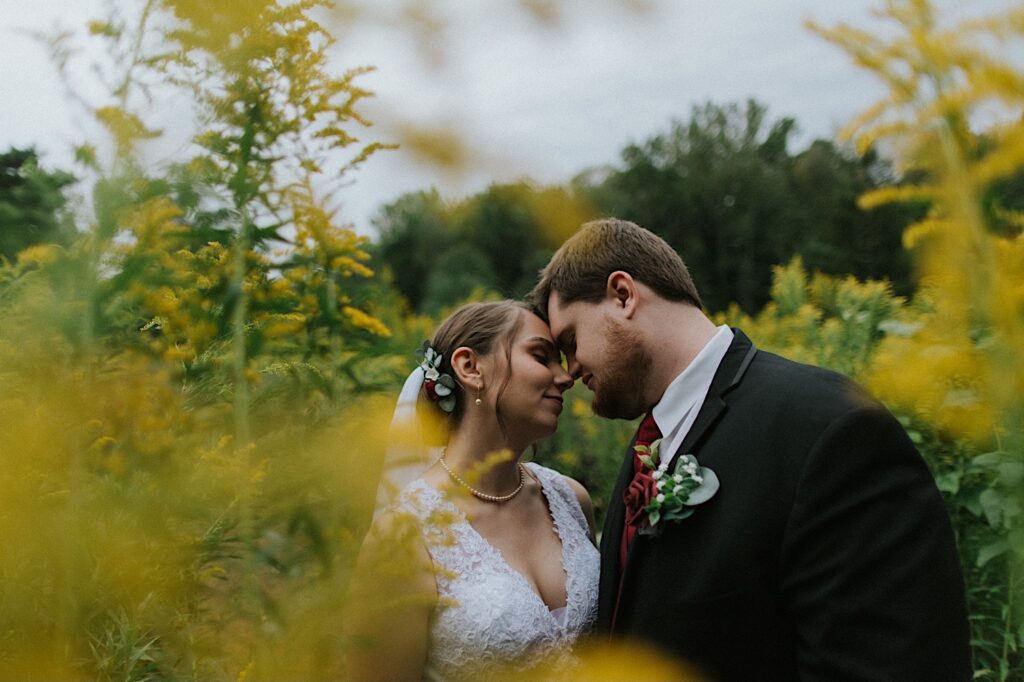 A bride and groom stand in a field of flowers together with their eyes closed and their noses touching with yellow flowers around them