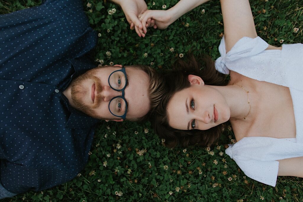 A man and a woman lay in the grass during their engagement session at Allerton Park and look up towards the camera while touching hands