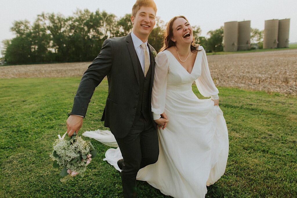 A bride and groom walk hand in hand in an empty field with some silos in the background as they laugh and smile at the camera