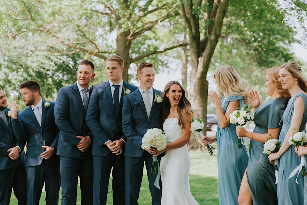 A bride smiles at her bridesmaids while standing next to her groom and her groomsmen in a park before their wedding ceremony at the Bloomington Country Club