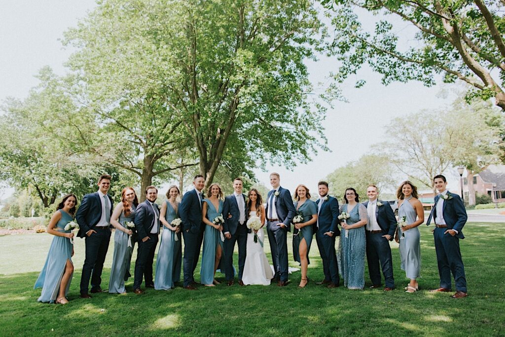 A bride and groom stand in a park with their wedding parties mixed on either side of them as they all smile at the camera before their wedding ceremony at Bloomington Country Club