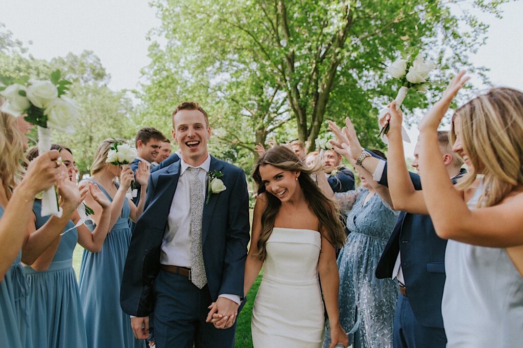 A bride and groom walk hand in hand towards the camera while smiling as their wedding parties on either side of them cheering them on before their wedding ceremony at Bloomington Country Club