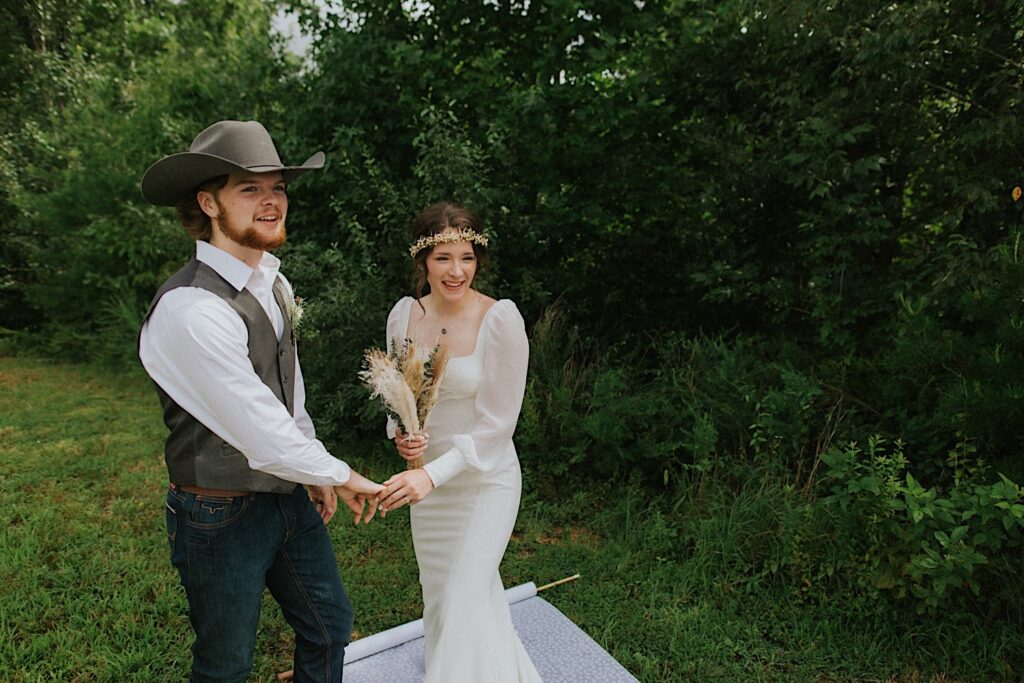 A bride and groom stand in front of a forest on a rug and touch hands while looking towards the camera and smiling