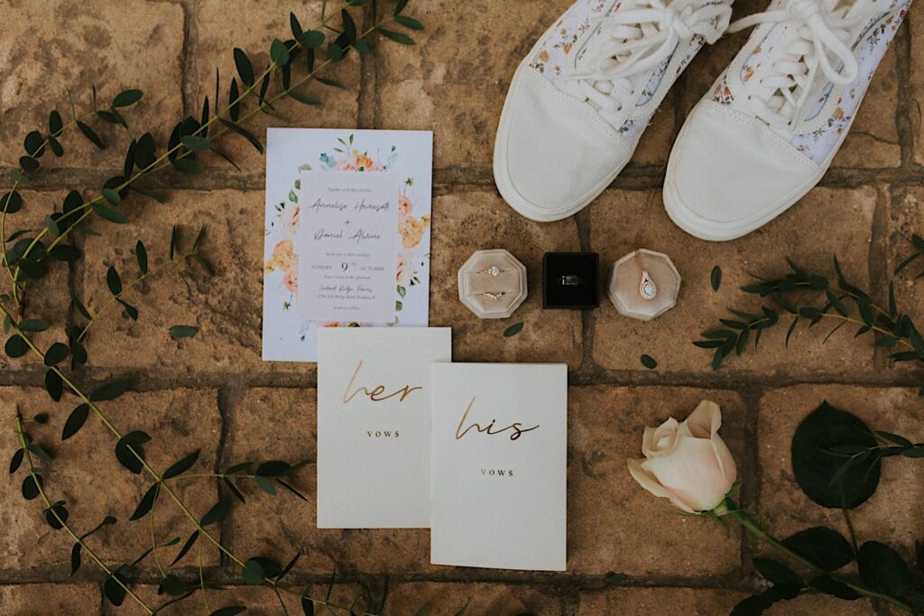 A wedding day flatlay with a brick background, included in the flatlay is an invite, vow books, wedding rings, shoes, a flower and other greenery