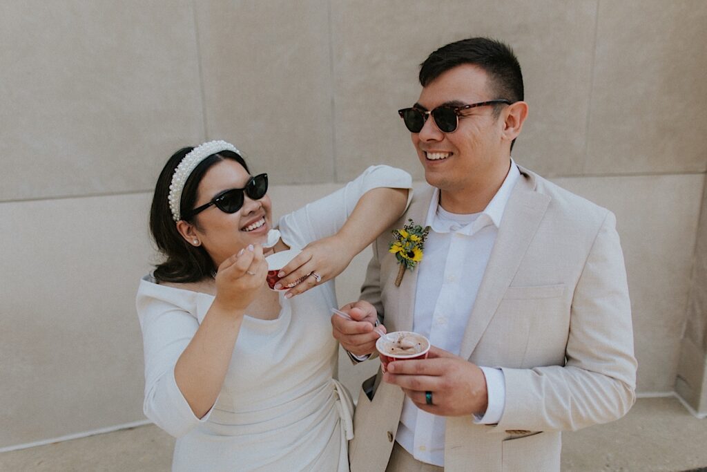A bride and groom with sunglasses on smile while standing next to one another in front of a stone wall while eating Cold Stone ice cream