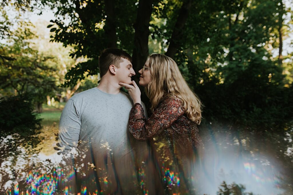 A man smiles as a woman touches his chin and leans in for a kiss while the two stand in a park during their engagement session in Springfield, Illinois