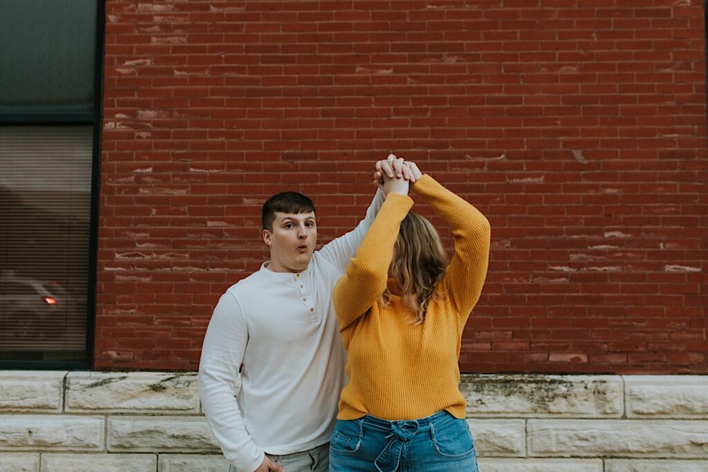 During an engagement session in downtown Springfield, Illinois a man makes a face at the camera while spinning a woman as they dance in front of a brick wall